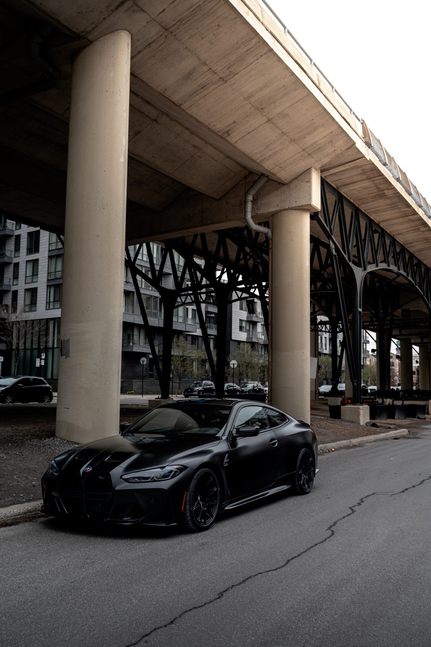 Matte black BMW M4 Competition beneath elevated structure, Montréal